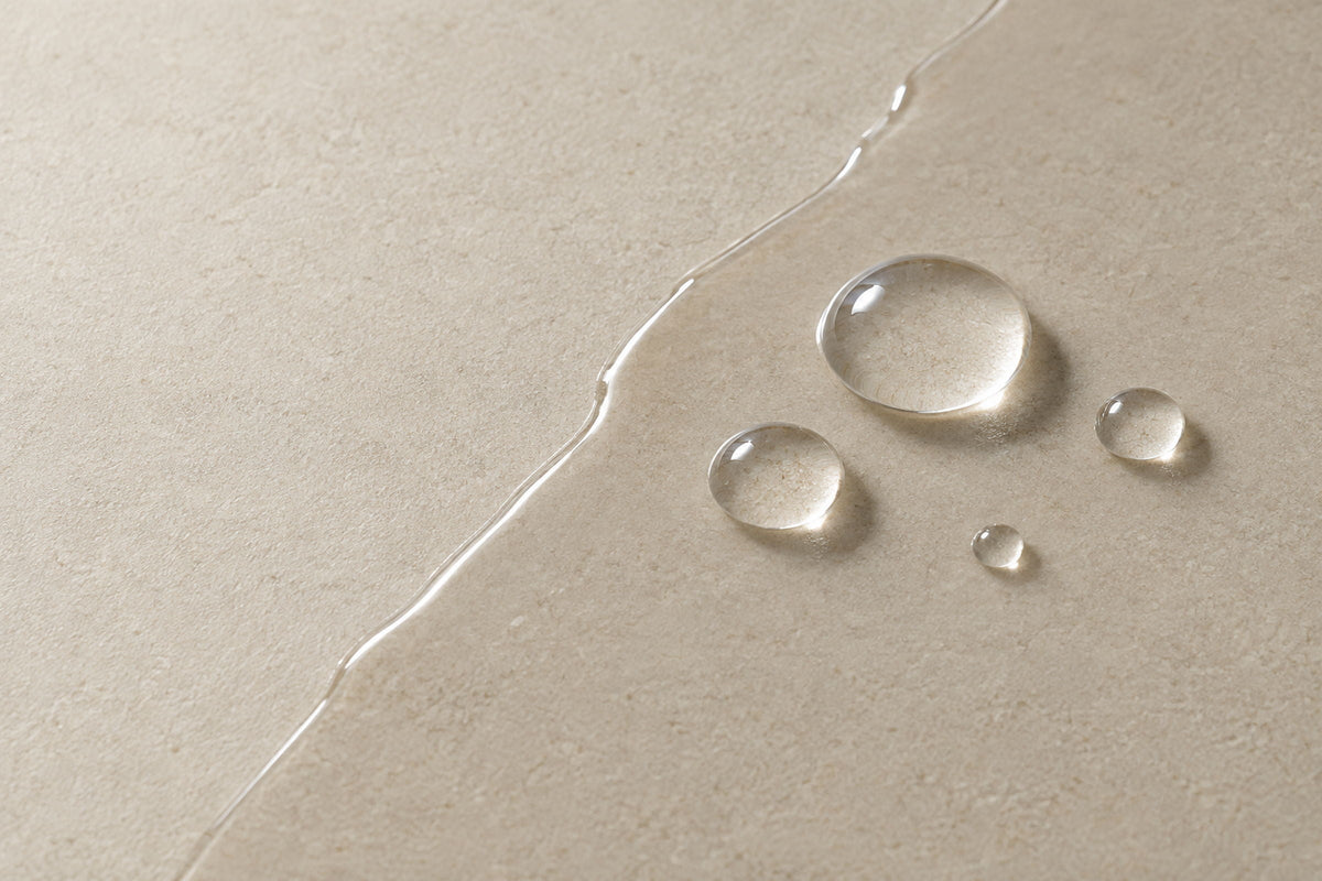 Four clear water droplets beaded on a thin oil film across a matte limestone surface, with the film's edge visible as a thin line.