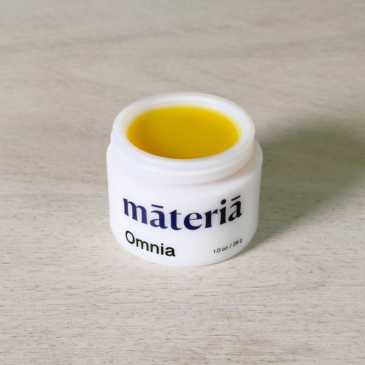 An open white cosmetic jar labeled 'Māteriā' and 'Omnia' with a bright golden yellow balm inside, placed on a light wood surface.