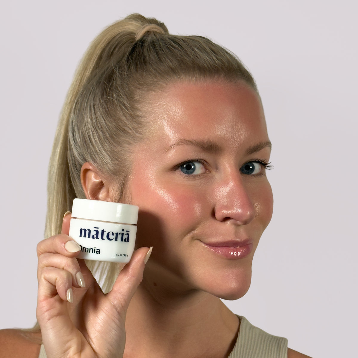 A woman with blonde hair in a high ponytail holds a white jar of Māteriā Omnia next to her cheek against a light gray studio background.
