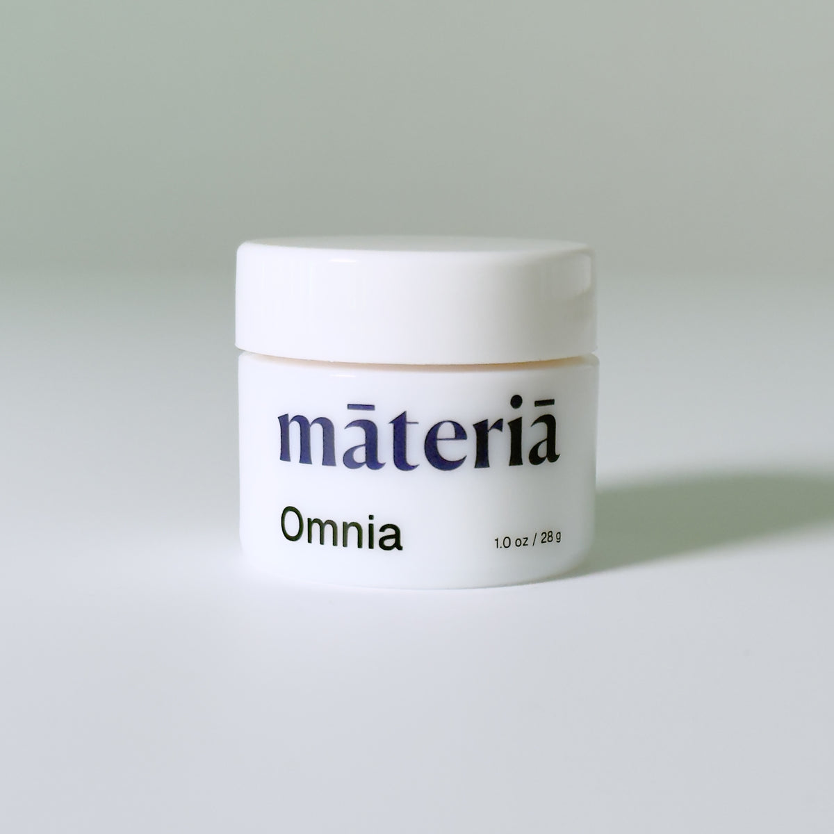 A white opal glass jar with a white lid, labeled 'Māteriā Omnia' in dark navy text, set against a clean, light gray background.