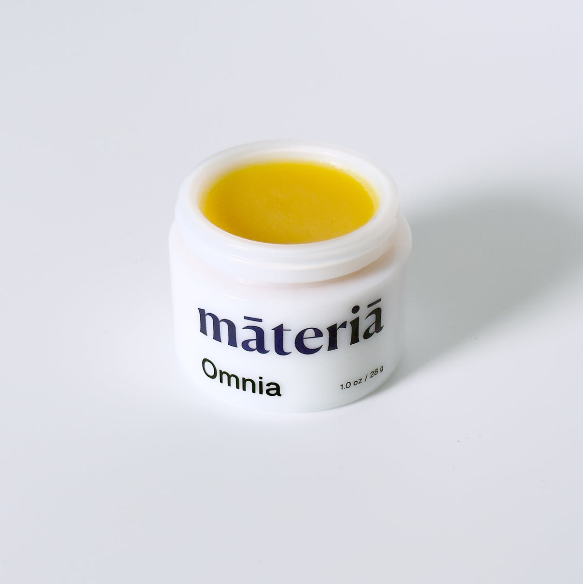 An open white opal glass jar of Māteriā Omnia balm, showing a bright yellow balm inside, set against a clean white background with a soft shadow.