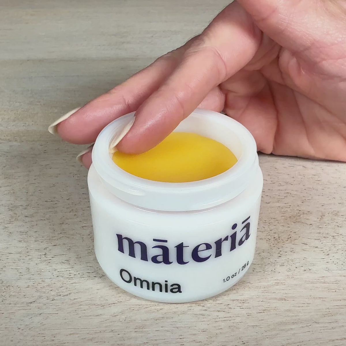 Short video of a fingertip pressing into the yellow balm inside an open white opal glass jar of Māteriā Omnia on a light wood surface.